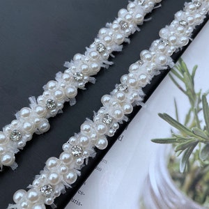 Puede incluir: Primer plano de dos hilos de adorno de perlas blancas con detalles de pedrer&iacute;a transparente. El adorno presenta peque&ntilde;as perlas redondas agrupadas, intercaladas con brillantes pedrer&iacute;a. El adorno se encuentra sobre un fondo oscuro.