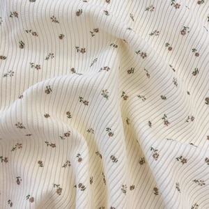 Può includere: Tessuto a coste color crema con un delicato motivo floreale. Il tessuto presenta coste verticali ed è adornato da piccoli disegni floreali marroni e verdi sparsi, creando un aspetto morbido e strutturato.