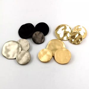 以下が含まれることがあります： さまざまなサイズと色の、不規則な形の金属製ボタンのコレクション。ボタンには、黒、銀、金の色合いがあり、テクスチャ加工されたハンマー加工の外観です。配置は、利用可能なさまざまな仕上げとサイズを示しています。