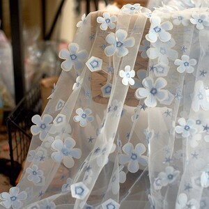May include: White tulle fabric with blue 3D flower embellishments. The fabric has a delicate, airy feel and is perfect for adding a touch of elegance to any project.