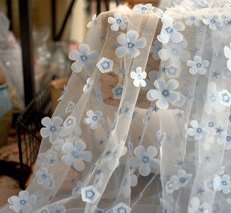May include: White tulle fabric with blue 3D flower embellishments. The fabric has a delicate, airy feel and is perfect for adding a touch of elegance to any project.