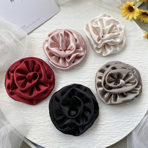 May include: Five circular hair scrunchies made of smooth, shiny satin fabric, each shaped like a blooming rose. The set includes colors in deep red, soft dusty pink, cream, black, and metallic taupe, all featuring gathered, layered fabric petals.