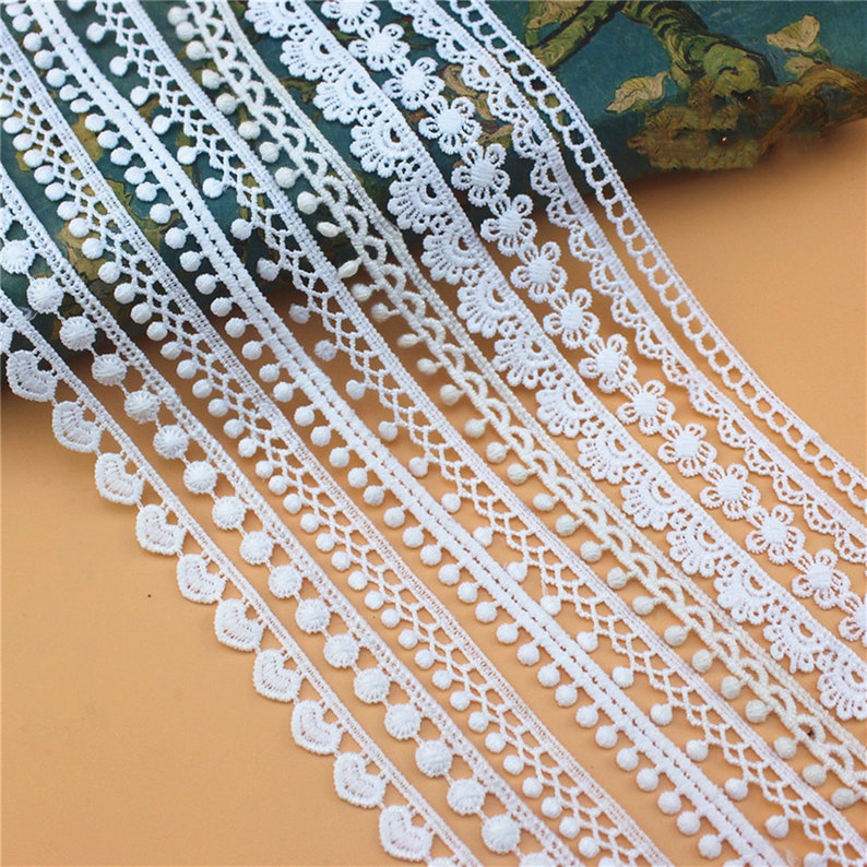 May include: A collection of white lace trims with various patterns, including scalloped edges, floral designs, and pom-poms. The trims are arranged in rows, showcasing their delicate details.
