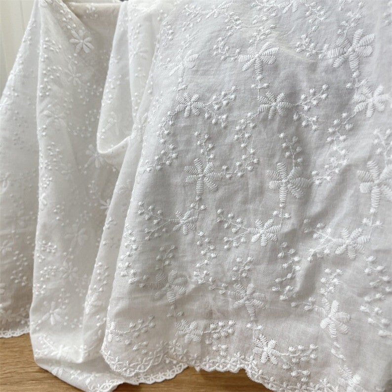 Puede incluir: Una tela blanca con un intrincado bordado floral en toda la superficie. El material muestra un patr&oacute;n repetitivo de peque&ntilde;as flores y delicadas enredaderas, terminado con un borde festoneado a lo largo del borde inferior del textil.
