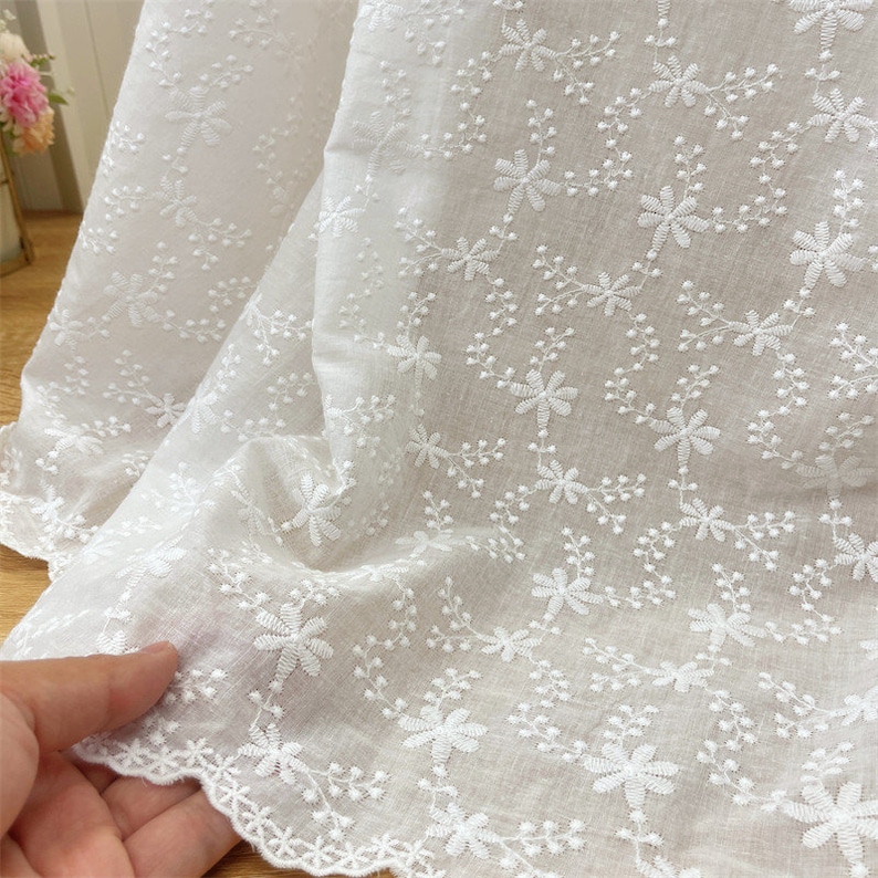 Puede incluir: Una tela blanca con un intrincado patr&oacute;n bordado de peque&ntilde;as flores y enredaderas delicadas. El material tiene una calidad semitransparente con un detalle de borde festoneado en la parte inferior, mostrando un motivo floral constante.