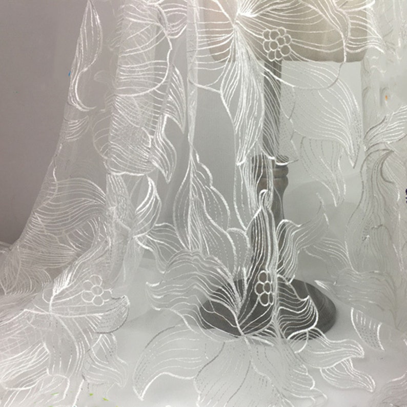 May include: White floral embroidered tulle fabric with a delicate, intricate design. The fabric is sheer and lightweight, making it ideal for a variety of crafting projects.