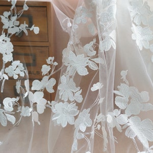 May include: Close-up of white floral lace fabric. The delicate lace features embroidered flowers and leaves, creating a textured pattern. The fabric is semi-sheer, with a soft, flowing drape. Ideal for bridal wear or decorative projects.
