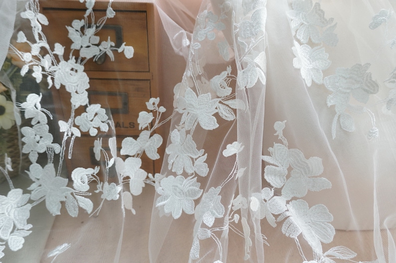 May include: Close-up of white floral lace fabric. The delicate lace features embroidered flowers and leaves, creating a textured pattern. The fabric is semi-sheer, with a soft, flowing drape. Ideal for bridal wear or decorative projects.