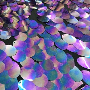 Puede incluir: Primer plano de lentejuelas iridiscentes en tonos morados, azules y plateados. Las lentejuelas de forma ovalada están dispuestas en un patrón superpuesto, creando una superficie texturizada. Cada lentejuela tiene un pequeño agujero en la parte superior. El efecto general es brillante y llamativo.