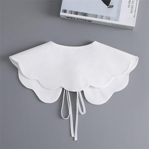 K&ouml;nnte beinhalten: Ein wei&szlig;er Kragen mit gewelltem Rand und mehrschichtigem Design. Der Kragen hat eine abgerundete Form und einen Bindebandverschluss vorne. Er besteht aus einem glatten, blickdichten Stoff auf grauem Hintergrund.