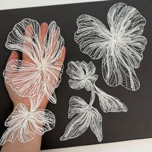 May include: Three white lace floral appliques with intricate, open-work mesh patterns. One large flower is positioned over a hand, while two additional floral motifs of varying sizes are displayed separately on a dark surface.