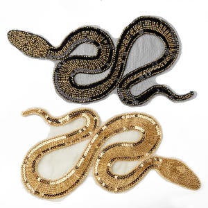 Puede incluir: Dos parches bordados con forma de serpiente. El parche superior presenta cuentas y lentejuelas negras y doradas sobre una base de malla negra. El parche inferior está elaborado con lentejuelas y cuentas doradas metálicas sobre malla clara.