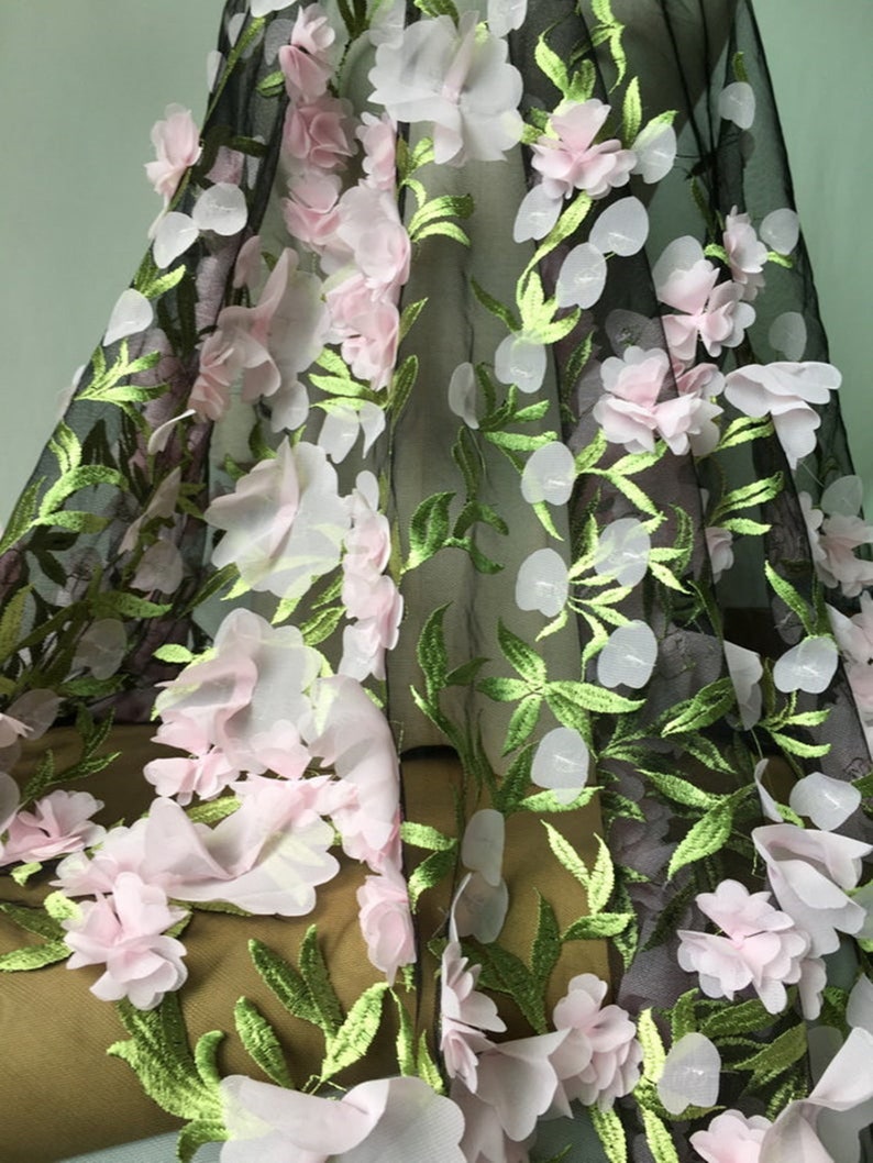 May include: Black mesh fabric with 3D pink and white flowers and green leaves. The fabric has a delicate and intricate design.