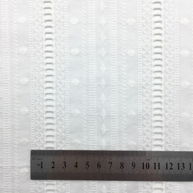 May include: White cotton fabric with a repeating embroidered pattern of small circles and geometric shapes. A ruler is placed at the bottom of the image, showing measurements in centimetres and millimetres.
