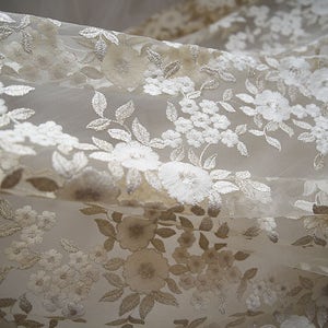 Pode incluir: Tecido de renda floral branca com um padrão delicado. A renda é transparente e tem uma textura macia e fluida.