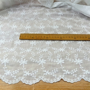 Puede incluir: Una tela blanca con un patr&oacute;n floral bordado de peque&ntilde;as flores y enredaderas. Una regla de madera est&aacute; colocada sobre el material, mostrando una escala de medici&oacute;n que llega hasta los 25 cent&iacute;metros, resaltando los detalles del bordado.