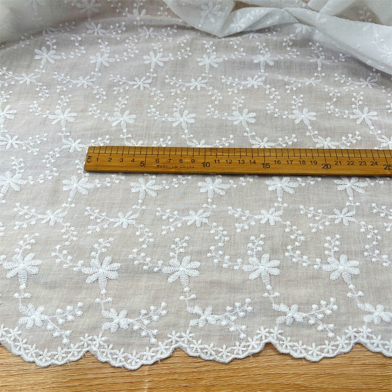 Puede incluir: Una tela blanca con un patr&oacute;n floral bordado de peque&ntilde;as flores y enredaderas. Una regla de madera est&aacute; colocada sobre el material, mostrando una escala de medici&oacute;n que llega hasta los 25 cent&iacute;metros, resaltando los detalles del bordado.