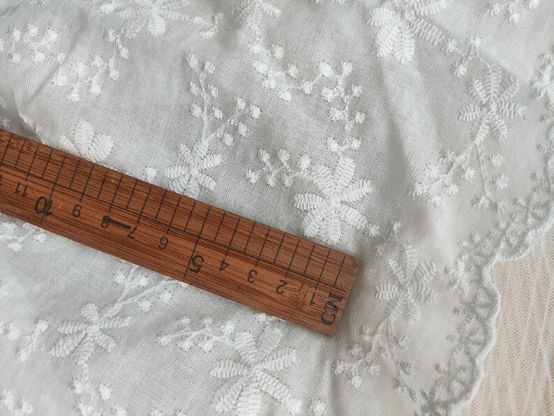 Puede incluir: Una tela blanca con intrincados bordados florales blancos y un borde festoneado. Una regla de madera est&aacute; colocada sobre el material, mostrando una escala de medici&oacute;n que supera los 28 cm, proporcionando una idea clara de la escala de la tela.