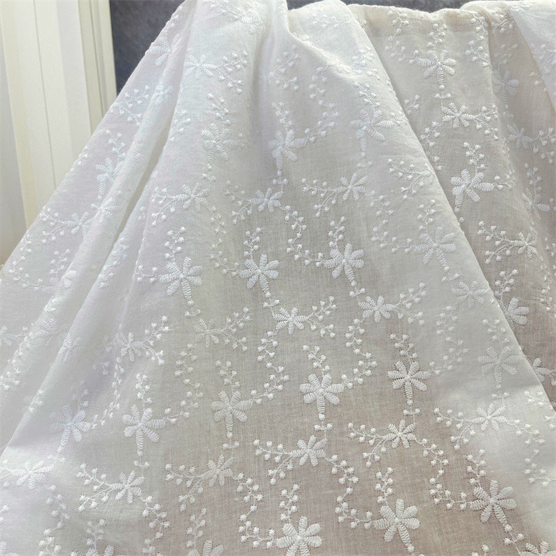 Puede incluir: Una tela blanca con un intrincado patr&oacute;n de bordado de peque&ntilde;as flores delicadas y enredaderas. El bordado est&aacute; realizado con hilo blanco, creando una textura tonal sutil sobre el material ligero y ligeramente transparente.
