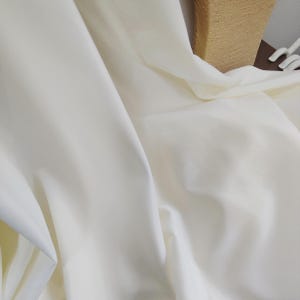 Puede incluir: Primer plano de una tela fluida de color crema, con pliegues suaves y sombras sutiles. La tela parece lisa y posiblemente sedosa, con un ligero brillo. Los pliegues crean una sensación de profundidad y textura, sugiriendo una cortina o prenda.