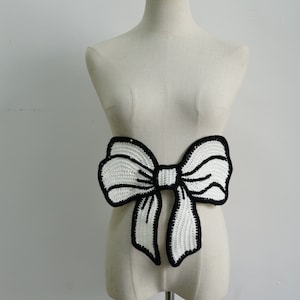 May include: A white crocheted bow with black outlining. The bow is large and has a 3D effect.