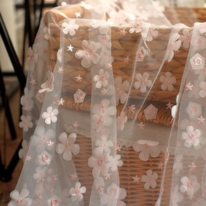 May include: White sheer fabric with pink 3D flower embellishments. The fabric is draped over a wicker basket.