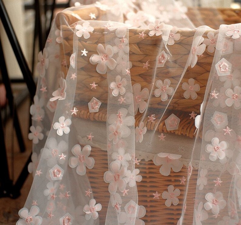 May include: White sheer fabric with pink 3D flower embellishments. The fabric is draped over a wicker basket.