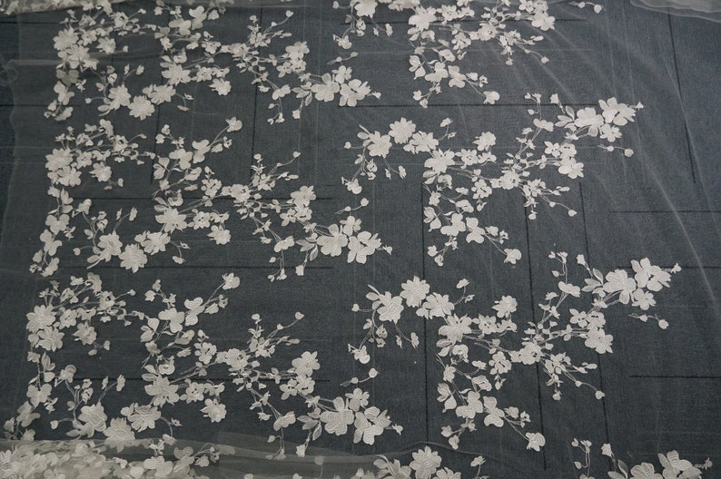May include: White floral lace fabric with a delicate, embroidered design. The pattern features clusters of flowers and leaves, creating a romantic and elegant aesthetic. Ideal for bridal wear or decorative projects.