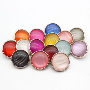 Può includere: Assortimento di 15 bottoni rotondi iridescenti con bordi in metallo dorato. I bottoni sono di vari colori, tra cui rosso, arancione, giallo, verde, blu, viola, rosa e bianco.