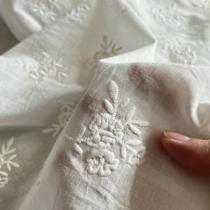 以下が含まれることがあります： 繊細な花の刺繍が施された白い生地のクローズアップ。刺繍は、花と葉の細かい模様が特徴です。生地はわずかに半透明で、折り目や質感が確認できます。全体的な美しさは柔らかくエレガントです。