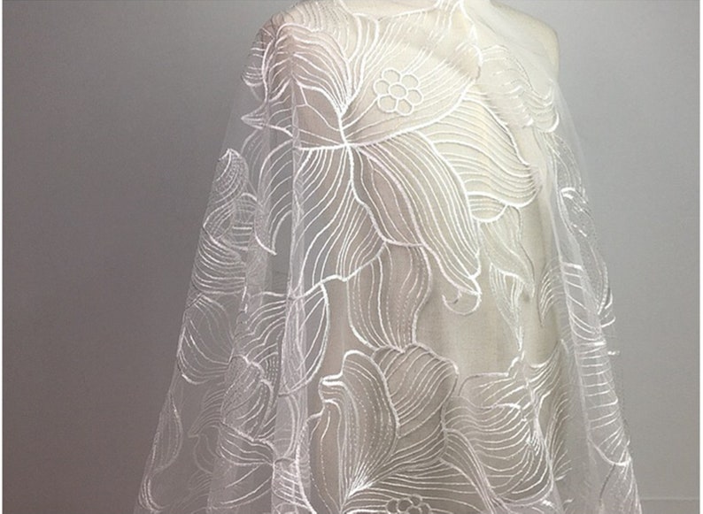 May include: White floral lace fabric with a delicate, intricate design. The lace is sheer and has a soft, flowing texture.