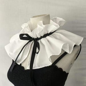 May include: A white ruffled collar with a black ribbon bow. The collar is layered and has a vintage aesthetic. The collar is shown on a mannequin over a black top.
