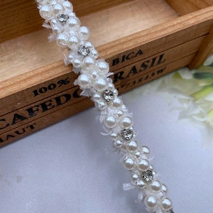 Puede incluir: Una diadema blanca adornada con perlas de imitaci&oacute;n y brillantes pedrer&iacute;a. Las perlas est&aacute;n agrupadas, creando un efecto texturizado. La diadema est&aacute; sobre un fondo de madera.