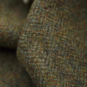 May include: Close-up of a textured fabric with a mix of green, brown, and blue threads. The material has a woven appearance, with visible fibers and a slightly rough texture. The fabric appears to be a tweed or similar woolen material.