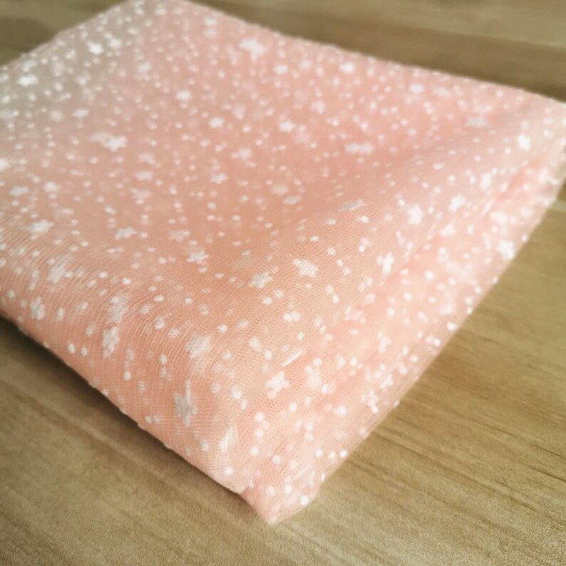 May include: Peach-colored tulle fabric with a white star pattern. The fabric is folded in half and has a soft, delicate texture.