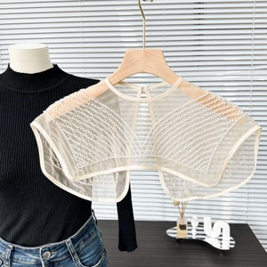May include: A sheer, off-white, beaded collar with a round neckline and short sleeves. The collar is decorated with rows of white pearls.