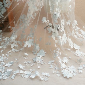 May include: Delicate white floral lace fabric with intricate embroidered flower and leaf details. The sheer material is ideal for bridal veils, dresses, or other decorative projects. The flowers are a soft white color.