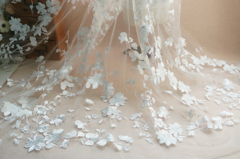 May include: Delicate white floral lace fabric with intricate embroidered flower and leaf details. The sheer material is ideal for bridal veils, dresses, or other decorative projects. The flowers are a soft white color.