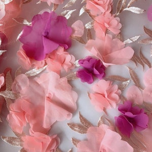 May include: Pink and purple 3D floral lace fabric with a delicate embroidered design. The fabric features a variety of flower shapes and sizes, with a soft, textured feel.