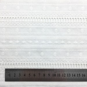 May include: White cotton fabric with an embroidered pattern of small circles, diamonds, and lines. A ruler is placed at the bottom of the image, showing measurements in centimetres.