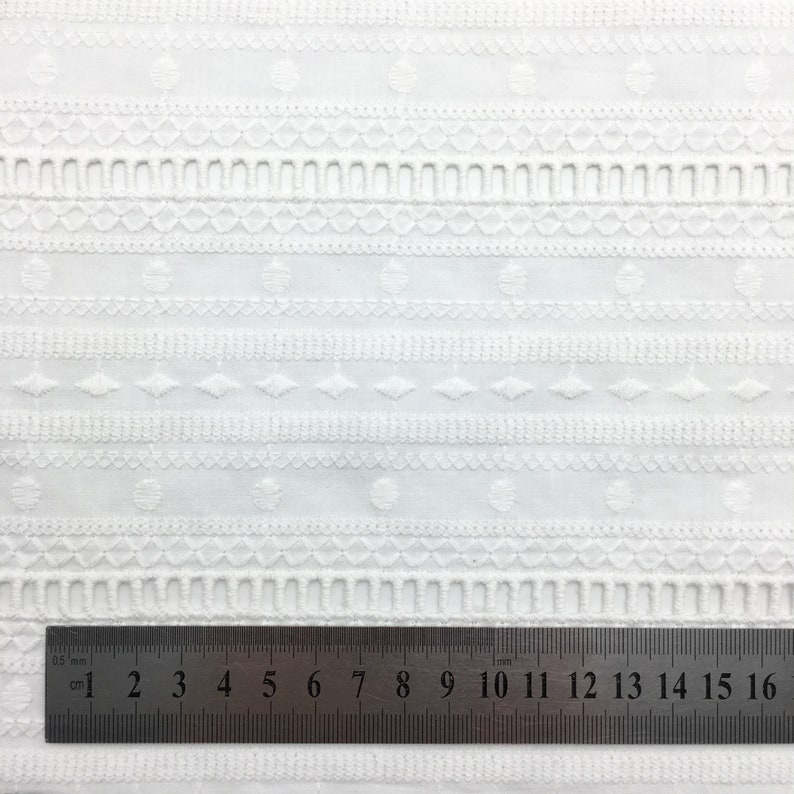 May include: White cotton fabric with an embroidered pattern of small circles, diamonds, and lines. A ruler is placed at the bottom of the image, showing measurements in centimetres.