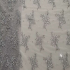 May include: A close-up of a white fabric with a silver floral pattern. The fabric is sheer and delicate, with a subtle shimmer.