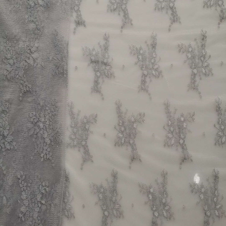 May include: A close-up of a white fabric with a silver floral pattern. The fabric is sheer and delicate, with a subtle shimmer.