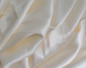 Puede incluir: Tela de satén blanco crema con una textura suave y sedosa. La tela está doblada y drapeada, mostrando su tacto lujoso.