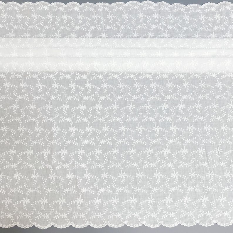 Puede incluir: Una tela blanca transparente con un intrincado patr&oacute;n bordado de peque&ntilde;as flores y delicadas enredaderas. El material tiene un borde festoneado en la parte superior e inferior, mostrando un motivo floral repetitivo en toda la superficie.