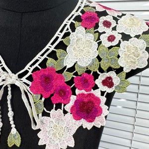 May include: White lace collar with pink and white floral embroidery. The collar has a tie fastening at the back.