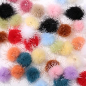 Puede incluir: Una colección de pompones coloridos y esponjosos en varios tonos, incluyendo negro, rosa, amarillo y azul. Cada pompón tiene una textura densa y suave con hebras largas y puntiagudas que se extienden hacia afuera. El surtido está disperso, creando una exhibición vibrante y juguetona.