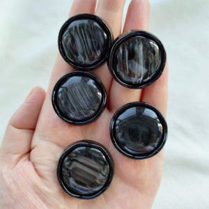 Może przedstawiać: Pięć okrągłych, czarnych kamieni obsydianu o wypolerowanej, odblaskowej powierzchni. Każdy kamień ma ciemny, wirujący wzór i jest osadzony w czarnej oprawie. Kamienie są prezentowane na jasnym tle.