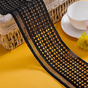 May include: A wide strip of black lace trim featuring a repeating geometric diamond pattern throughout the center and a linear, ribbed border along the edges. The lace is constructed with a fine mesh structure and distinct, dark thread work.
