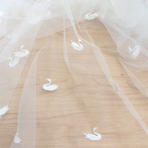 Peut inclure: Tissu de tulle blanc avec des motifs de cygnes brodés. Le tissu est transparent et délicat, avec une texture douce et fluide.